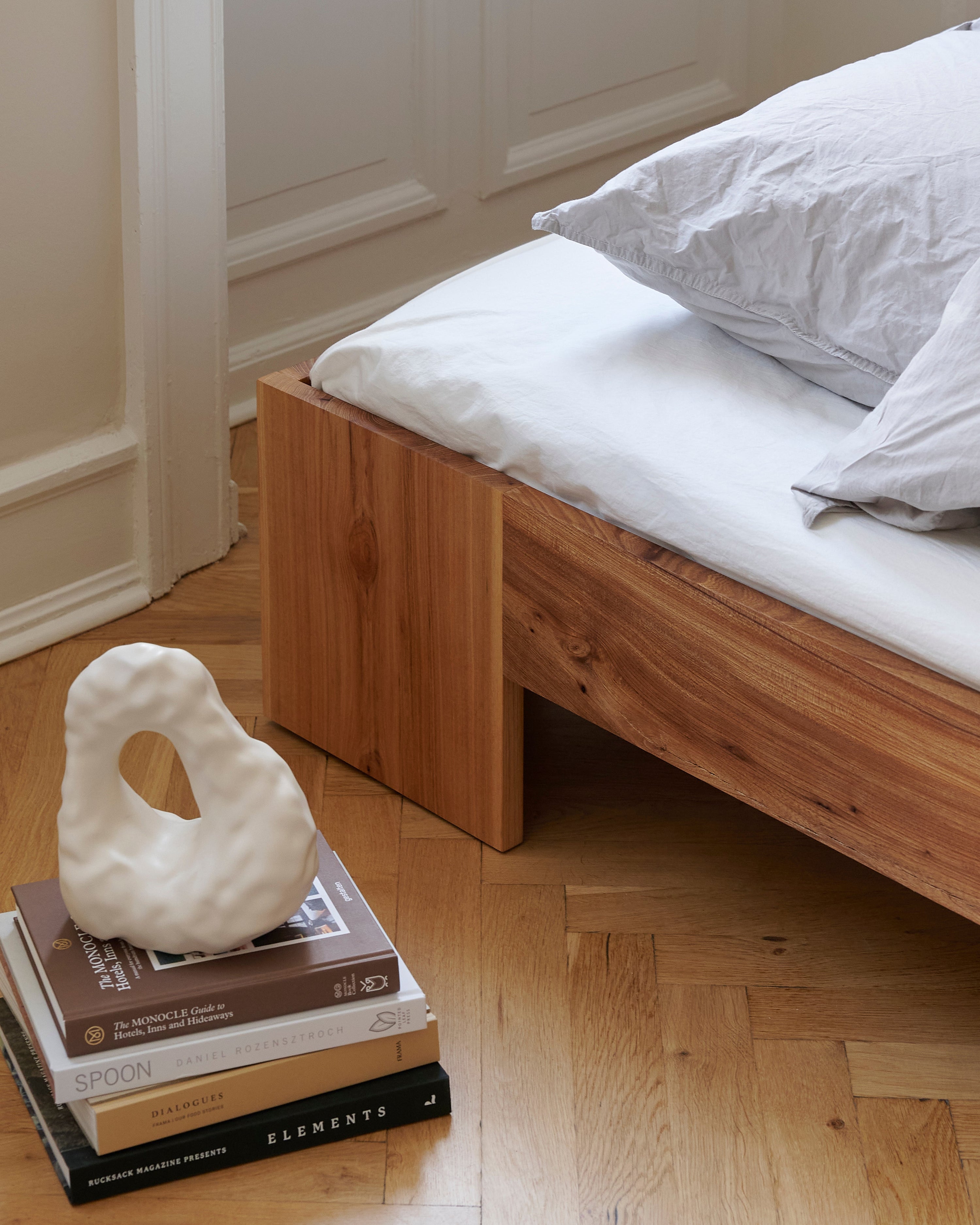 Wooden bed with white mattress and pillows, accompanied by a white abstract sculpture on books.